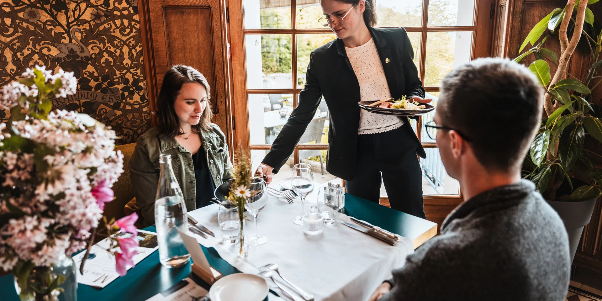 Un moment convivial dans un restaurant élégant et chaleureux. Une serveuse élégamment vêtue sert un plat raffiné à une table où un homme et une femme profitent d'un repas. La table est dressée avec soin, ornée de fleurs fraîches, de verres à pied et de menus. La lumière naturelle illumine la scène à travers de grandes fenêtres en bois, offrant une vue sur un jardin extérieur verdoyant.